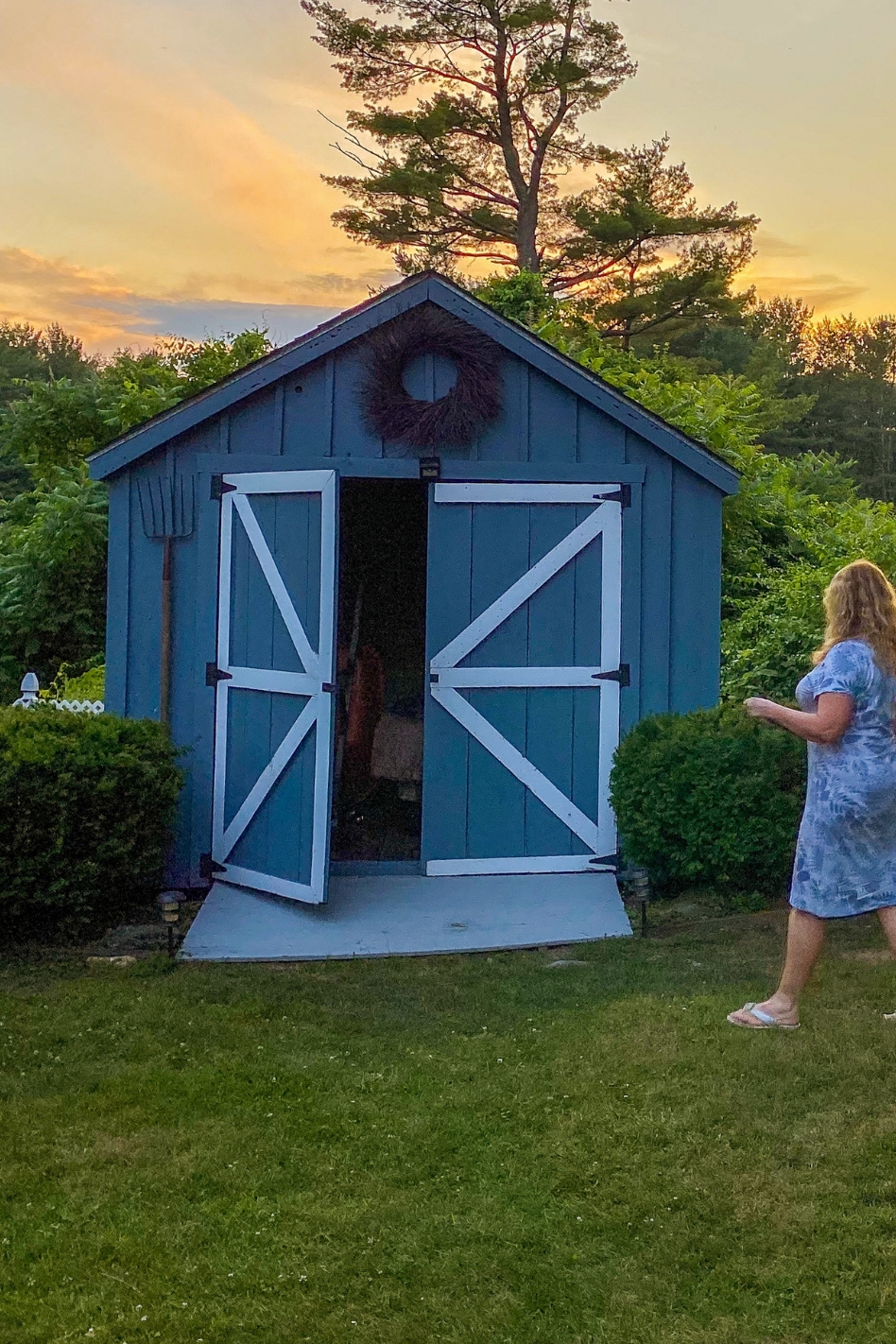 A woman walks on lawn towards an open entrance of a blue accessory building.