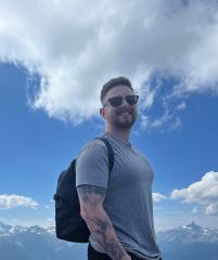 profile photo of man in gray shirt with mountains and blue sky in the background